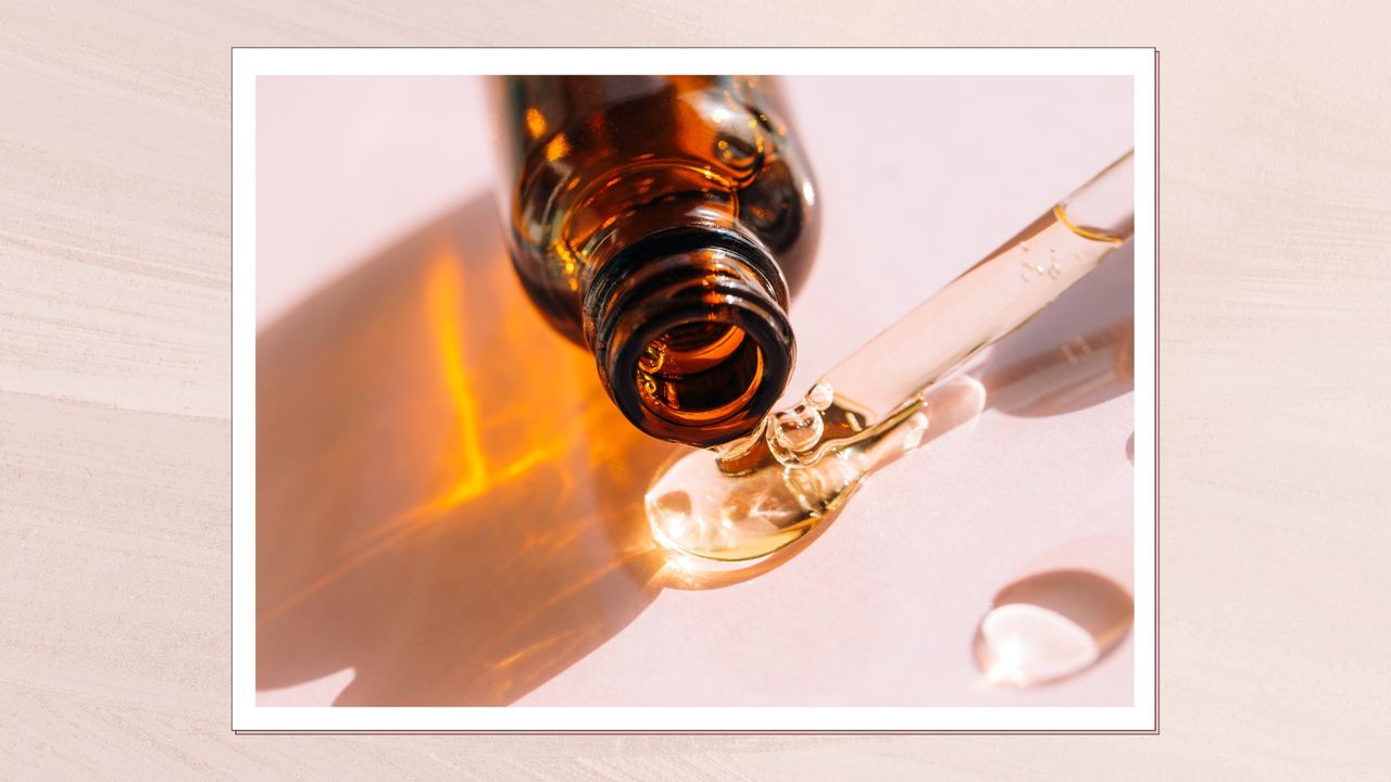 A close up of a brown glass serum bottle, lying on it&#039;s side, alongside a glass pipette with clear liquid coming out, on a pink backdrop/ in a pale pink textured template