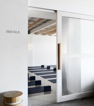 Partial view of a yoga studio through an open white and semi-transparent sliding door at Studio Warrior One. The studio features white walls, light wood ceiling beams, a wall light and multiple black yoga mats laid out on the floor. The word 'INDRA' can be seen on the wall outside the studio