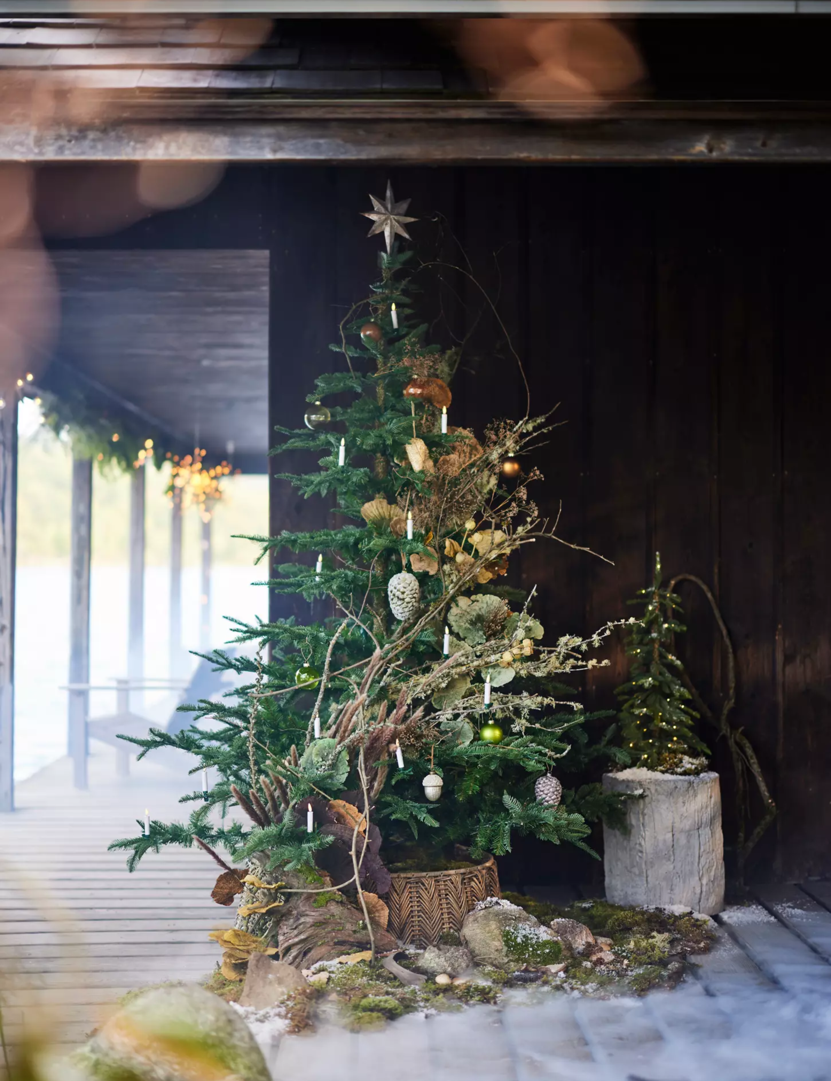 A Christmas tree styled before a rustic wooden cabin with clip-on candle ornaments, flocked pinecones, and metallic accents.