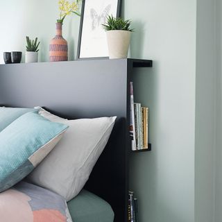a bedroom with mint green walls, the bed with a black headboard that has shelving storage behind it containing books, and a flat shelf on top holding a selection of vases