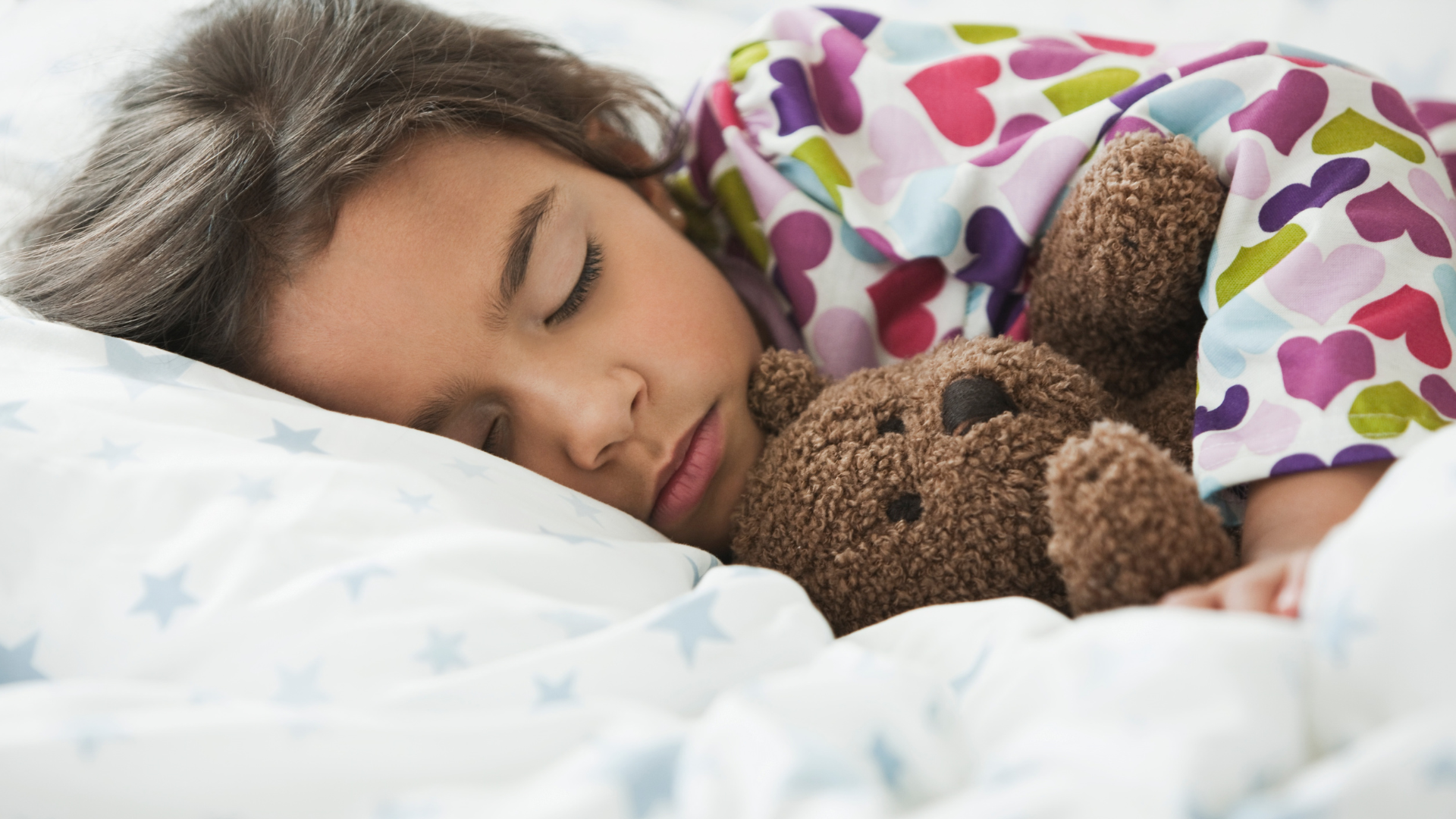 A slightly older child sleeps in a bed with star patterned bedding as she holds a brown teddy bear.