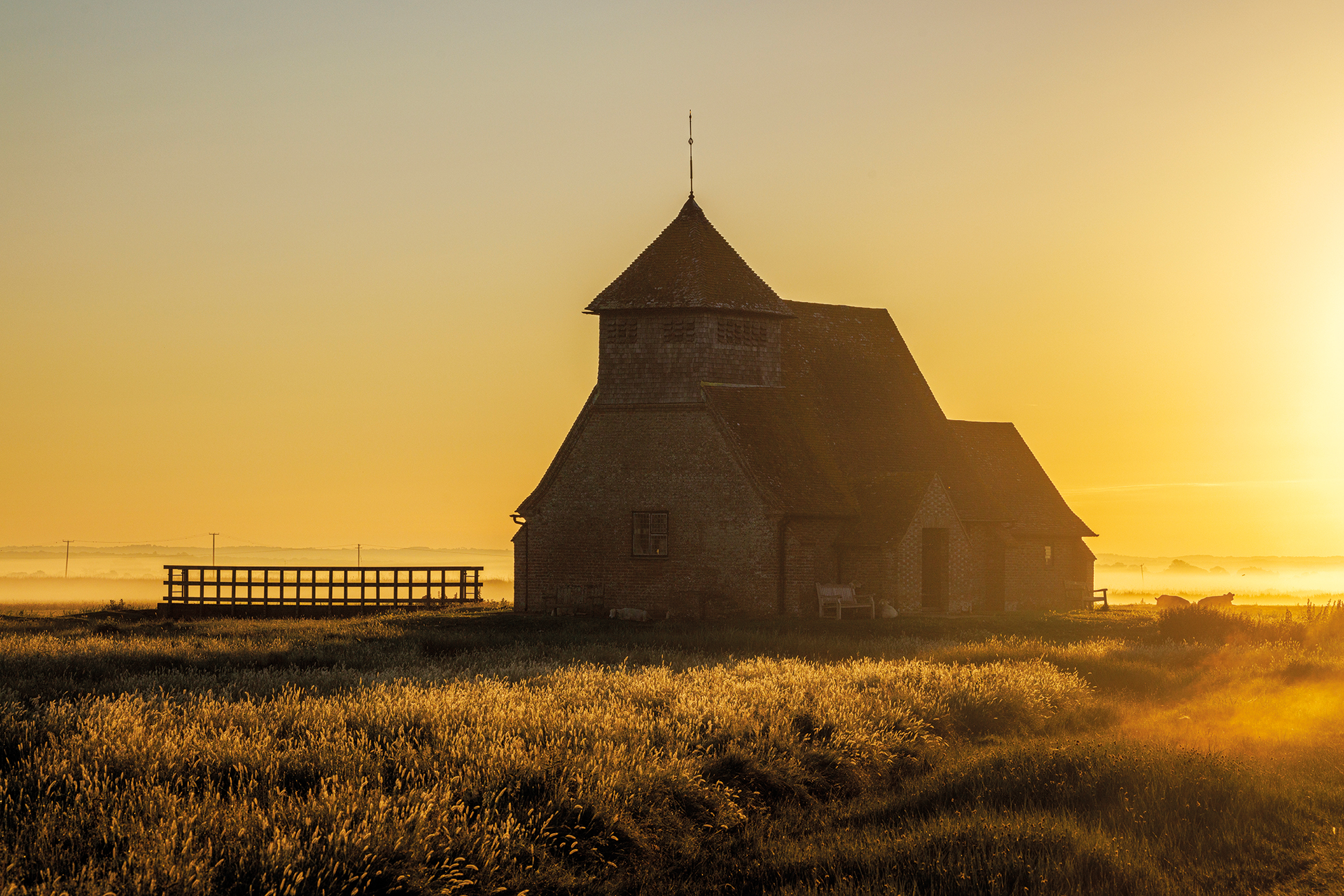 A rustic church stands silhouetted against a golden sunrise. Mist blankets the ground, creating a serene and peaceful rural landscape