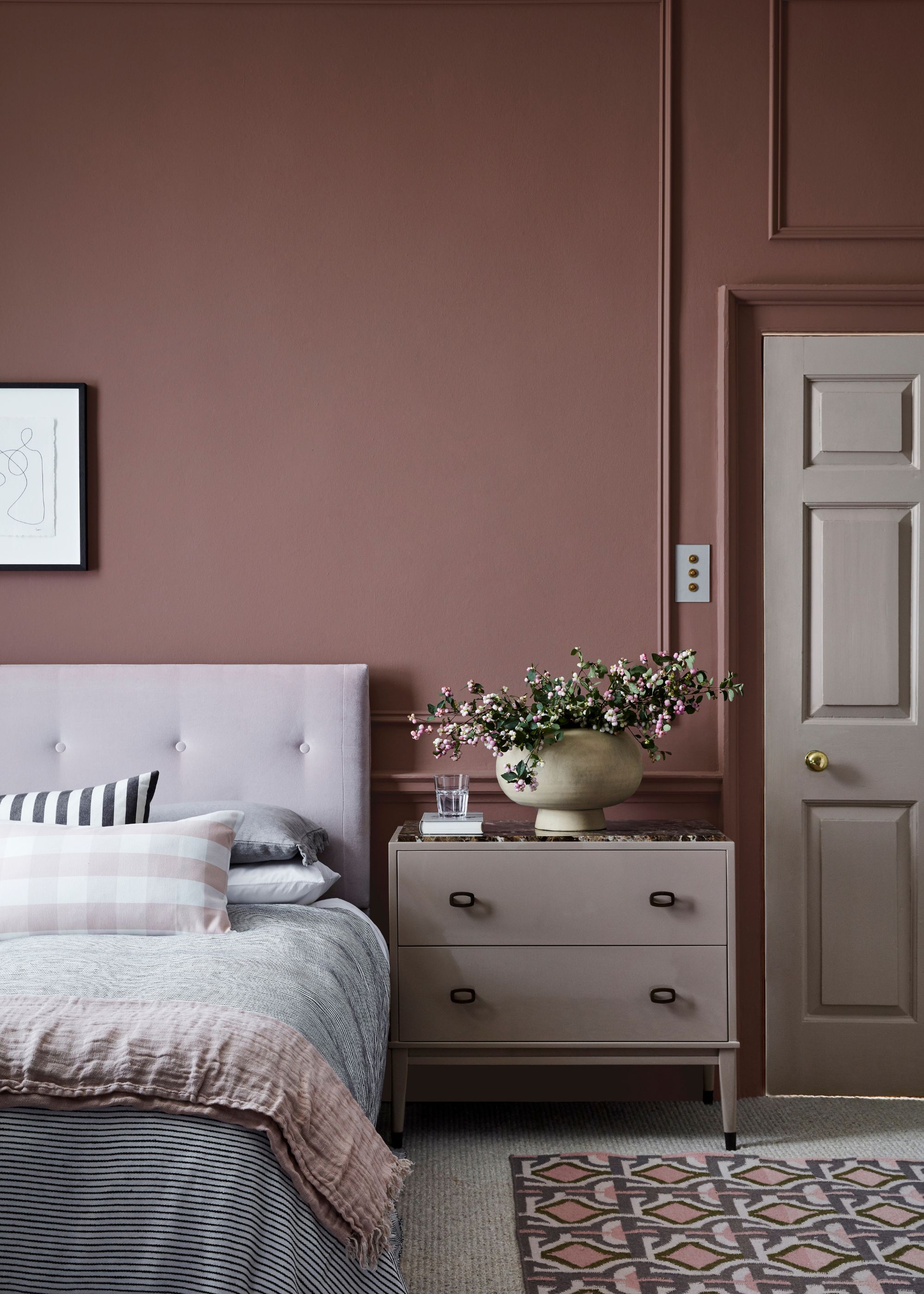 bedroom with dusky pink walls, light gray fabric headboard, patterned rug and taupe nightstand with vase of flowers