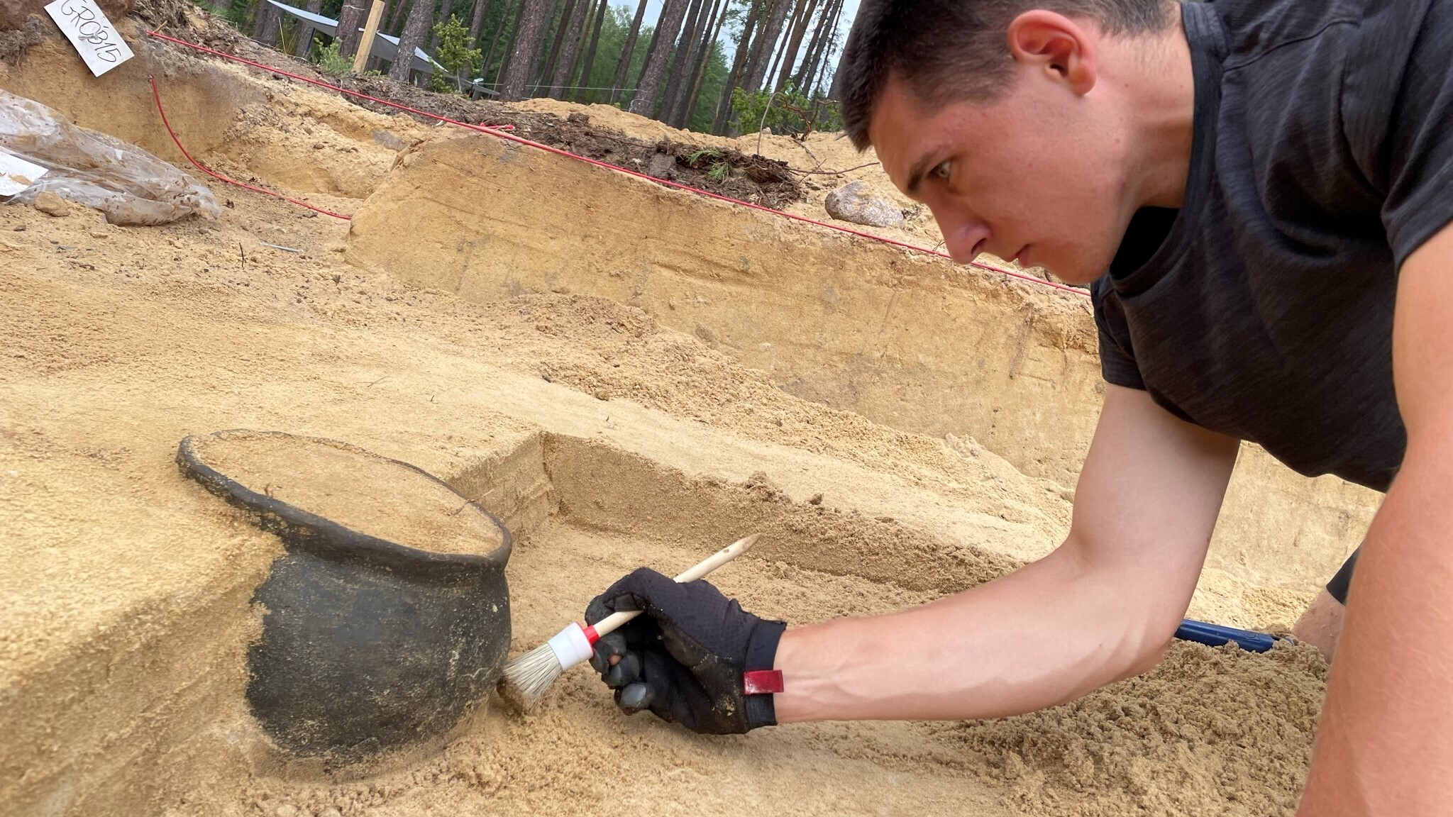 A researcher excavates a black cremation urn in a dirt-filled excavation space.