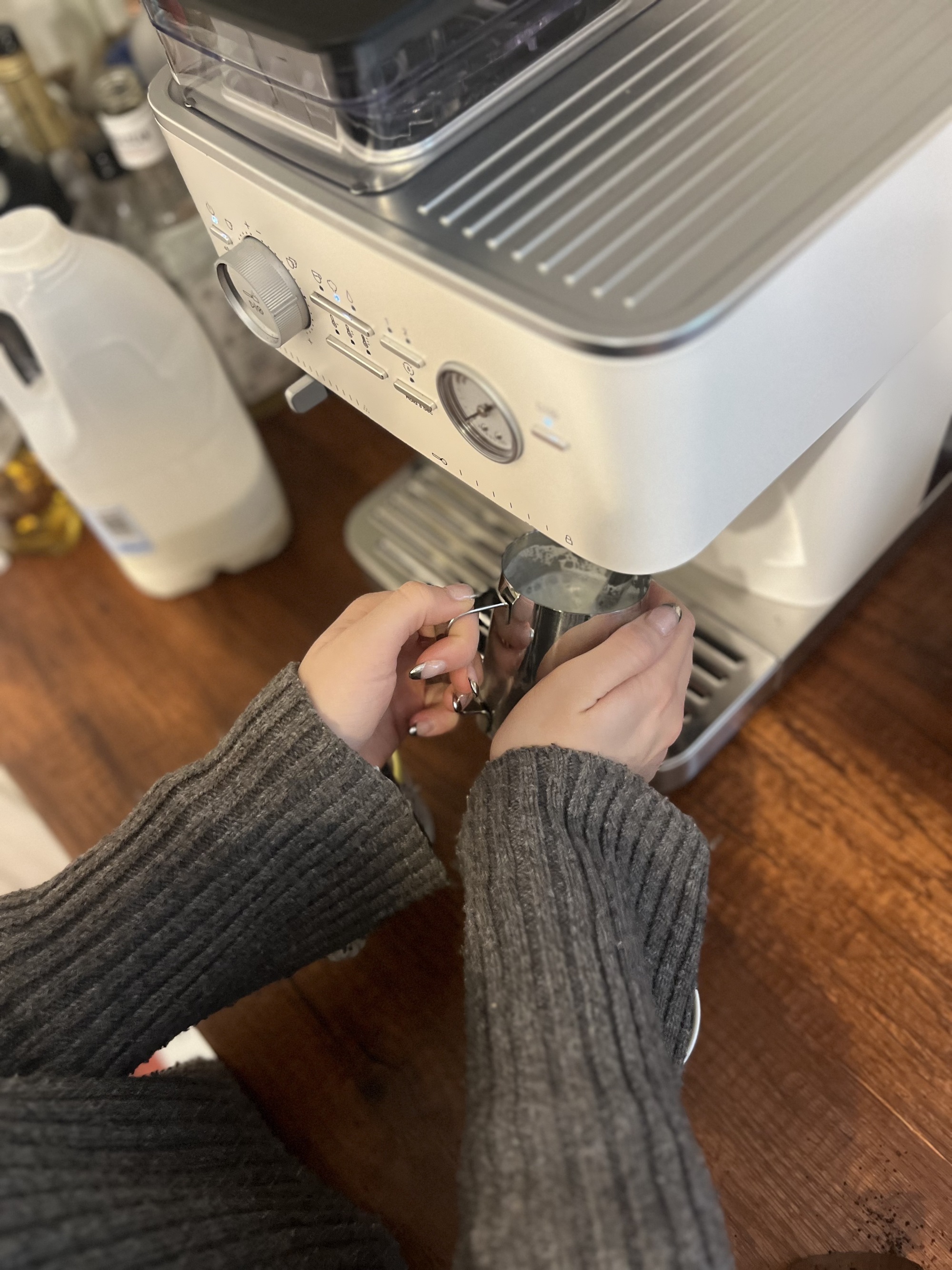 Image of someone wearing a gray sweater steaming milk by a steaming wand connected to a white espresso machine.