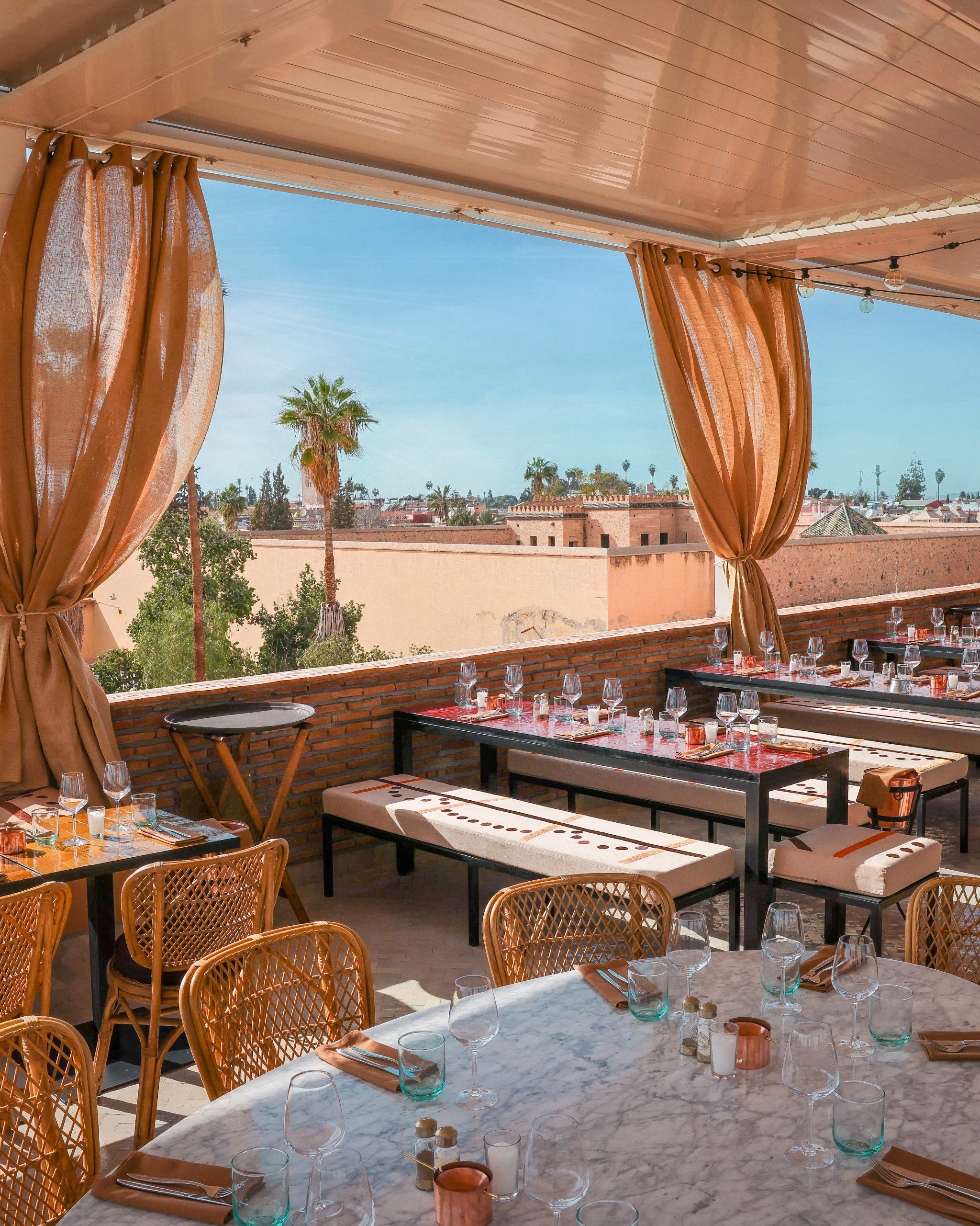 Rooftop restaurant terrace with rattan furniture and peach curtains floating in the wind against the blue sky.