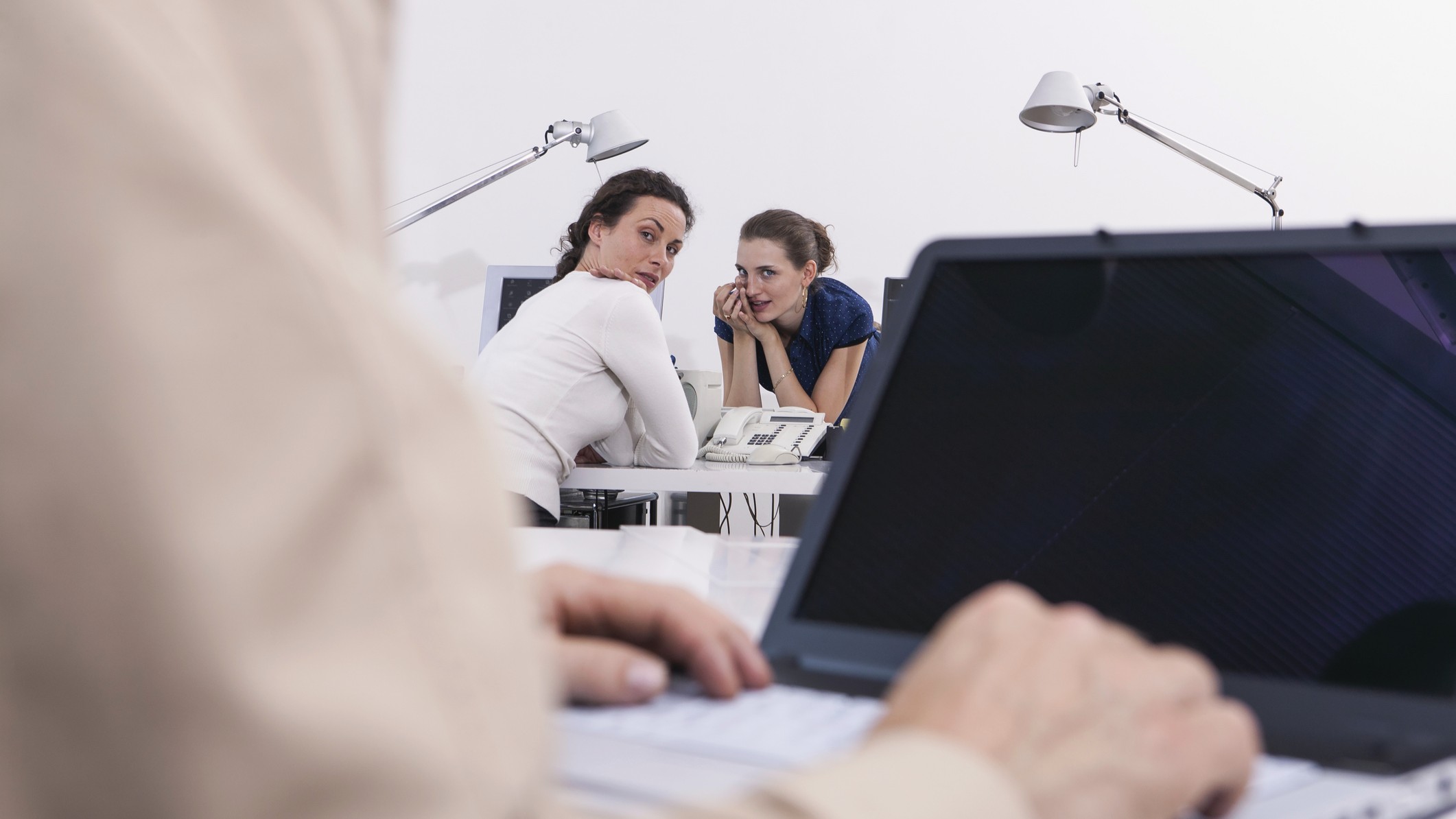 Two women in an office gossip with their heads together, looking at an older person in the foreground. We only see his laptop and hands.