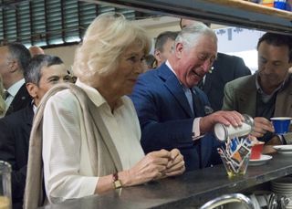 King Charles sitting at a counter and wearing a blue suit and tie dumping a glass jar of sugar into his red coffee cup at a counter surrounded by Queen Camilla and people behind them