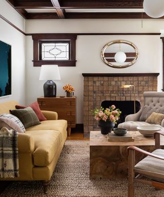 Traditional living room with dark, wood paneled ceiling, brick fireplace, yellow sofa, wooden block coffee table, small window with dark wooden frame, textured natural rug
