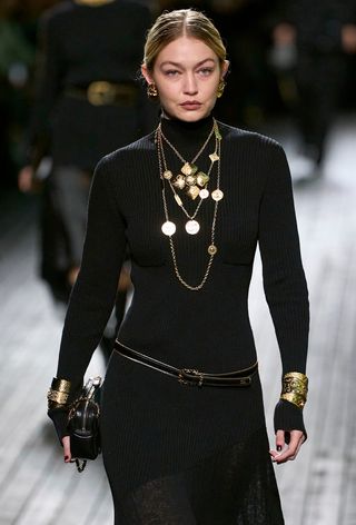 one of the best chunky cuff bracelets is shown in a photo from Chanel's fall 2024 runway show with a model wearing a black turtleneck dress with layered gold necklaces, a thin black belt, and stacked gold bangles