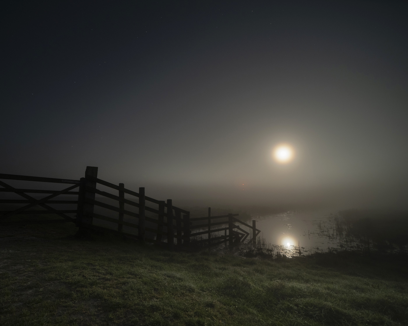 A misty moonlit night over the marsh, with a wooden gate and fence silhouetted beside a still pool reflecting the hazy moon.