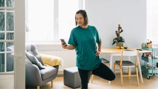 Woman standing on one leg wearing workout clothes in living room holding phone