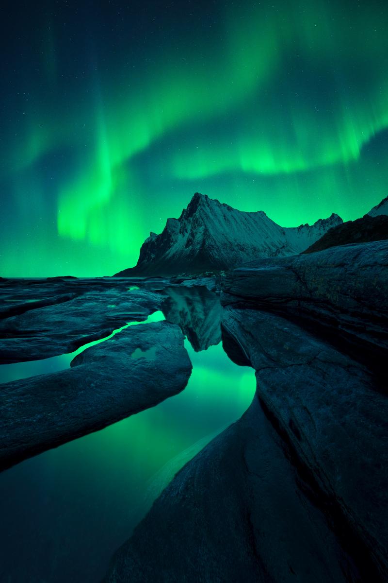 Green waves of aurora borealis streak the sky above a pointed, rocky mountain, rooted to the valley in the foreground with flowing streams that reflect the green auroras above.