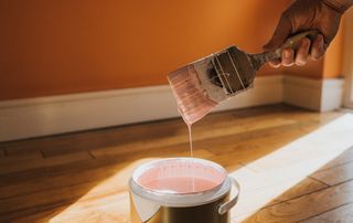 hand holding paintbrush with pink paint above paint tin, skirting board and floor in background