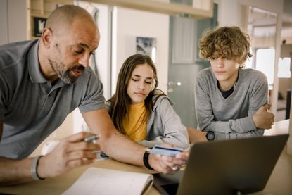 Brother and sister looking at father explaining finance with credit card at kitchen island 