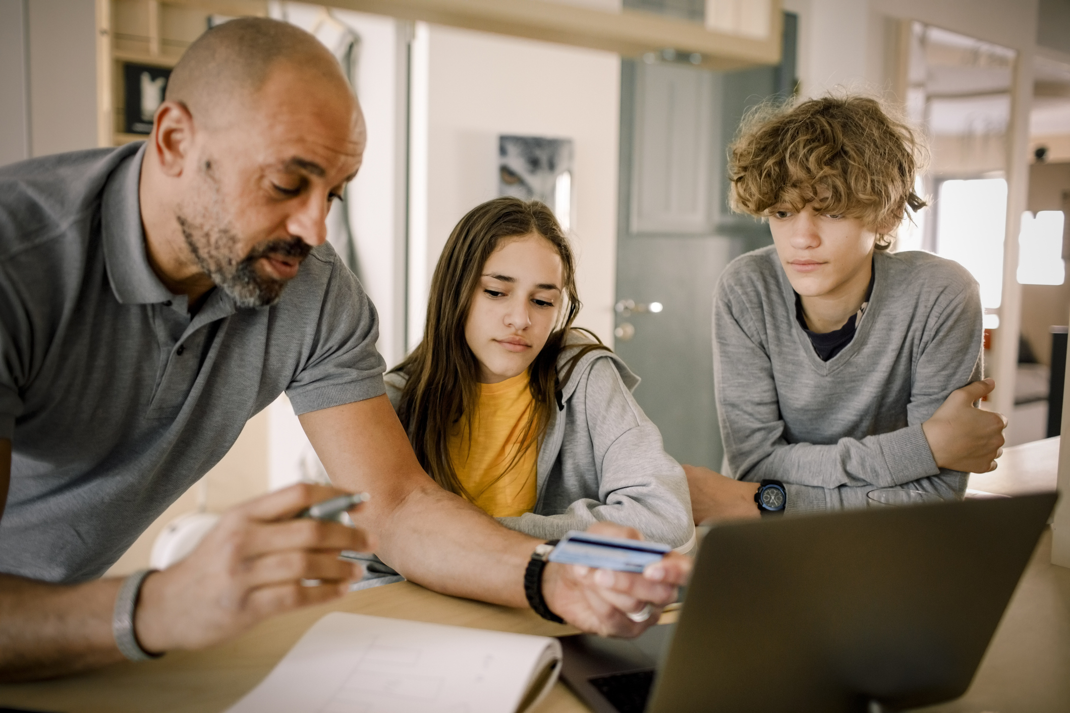Brother and sister looking at father explaining finance with credit card at kitchen island 