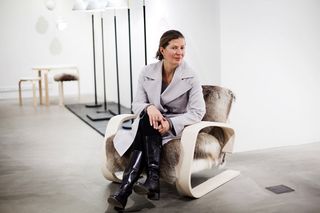 Ilse Crawford sitting on an Upholstered chairs in swedish reindeer fur. Behind her are 5 floor lights with black base and white shades. phoographed against a white wall