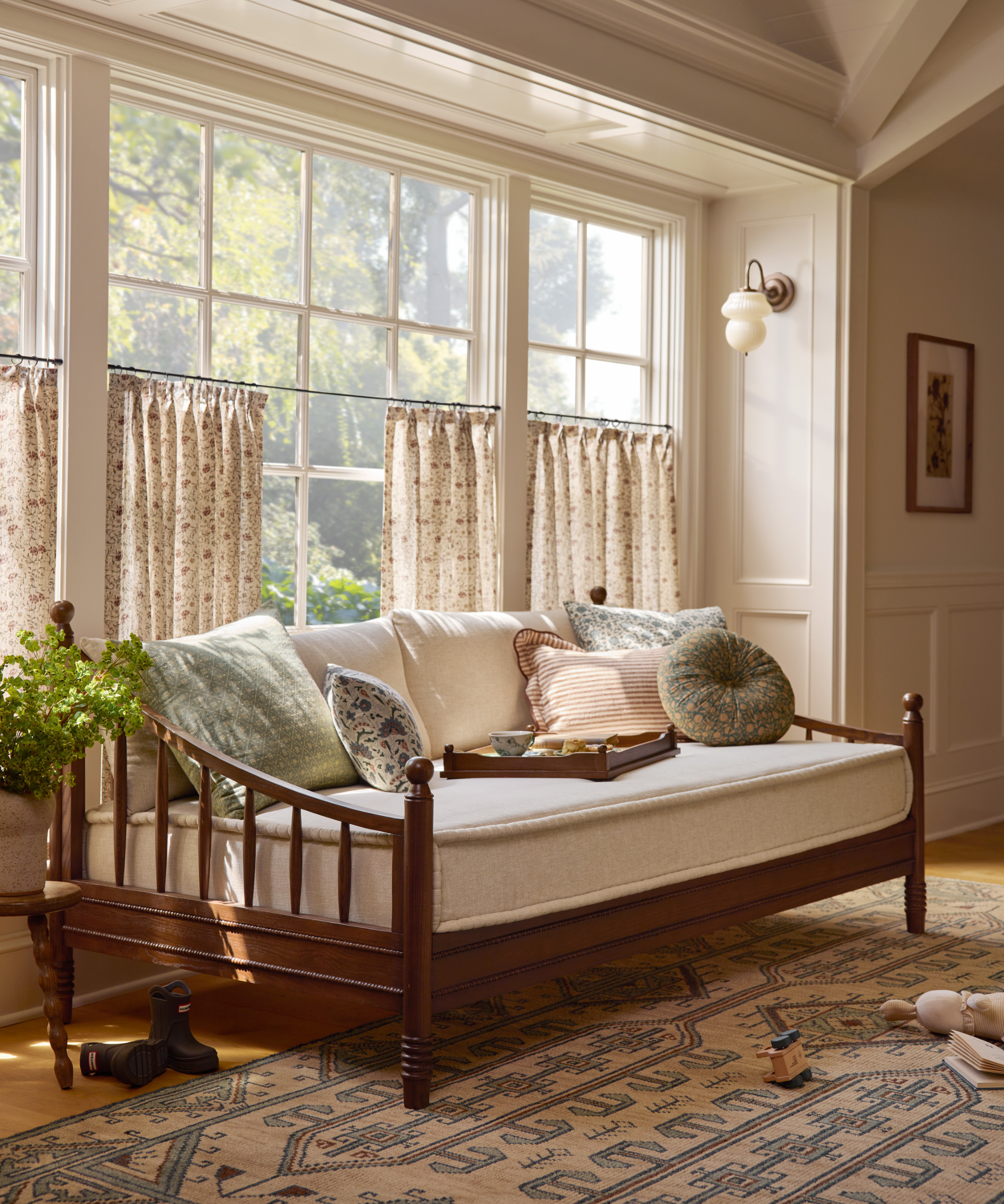 a wooden daybed with a tan seat cushion, lots of pillows, a geometric rug, and cafe curtains along the windows