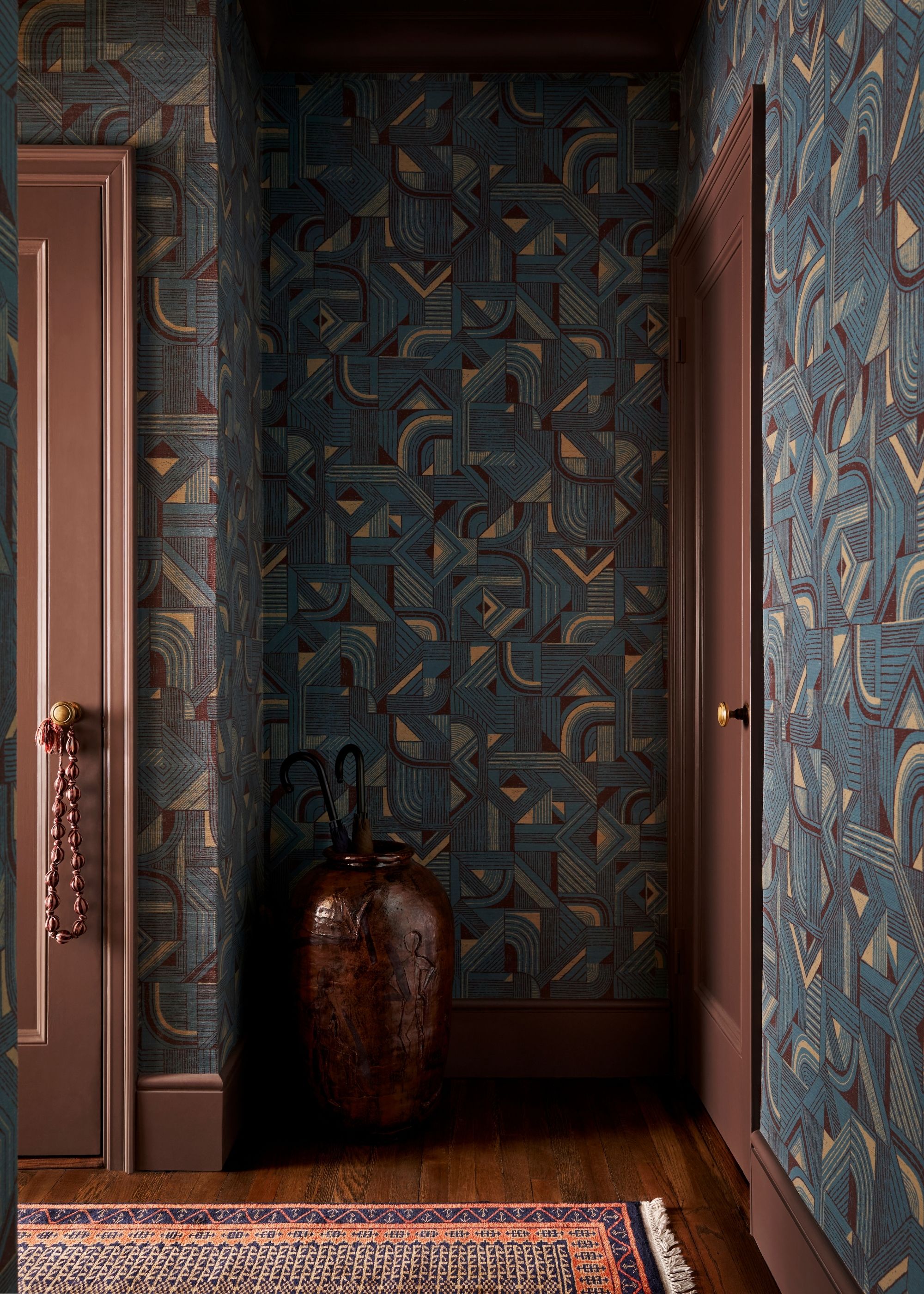 A dark and moody corner of a room with dark blue and purple patterned wallpaper on all walls, dark brown trim and doors, and wooden flooring.