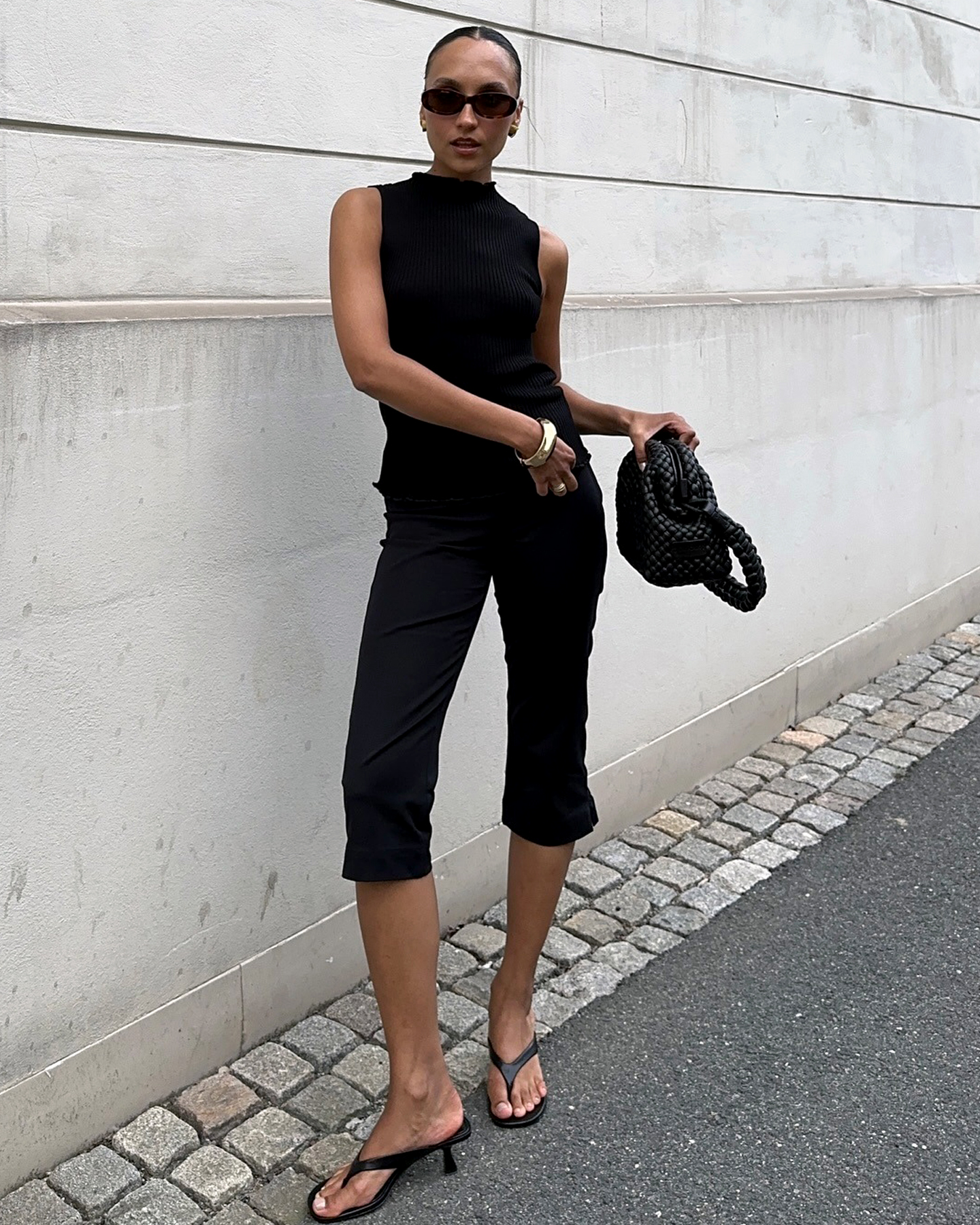 Style influencer Ingrid Fagerli Edvinsen poses in a chic all-black summer 2026 outfit with oval sunglasses, a mock-neck sleeveless top, woven bag, capri pants, and kitten-heel flip-flops.