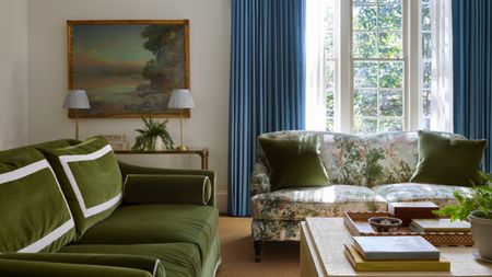 Living room with blue drapes and large French door, floral sofa with green cushions and a green velvet sofa, wooden coffee table with books and decor and an antique landscape above a console in the corner
