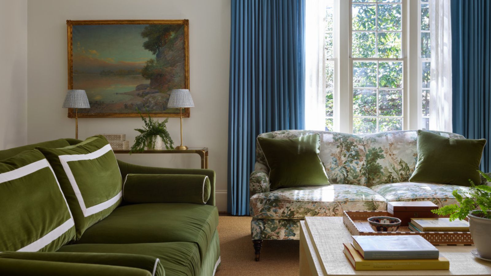 Living room with blue drapes and large French door, floral sofa with green cushions and a green velvet sofa, wooden coffee table with books and decor and an antique landscape above a console in the corner