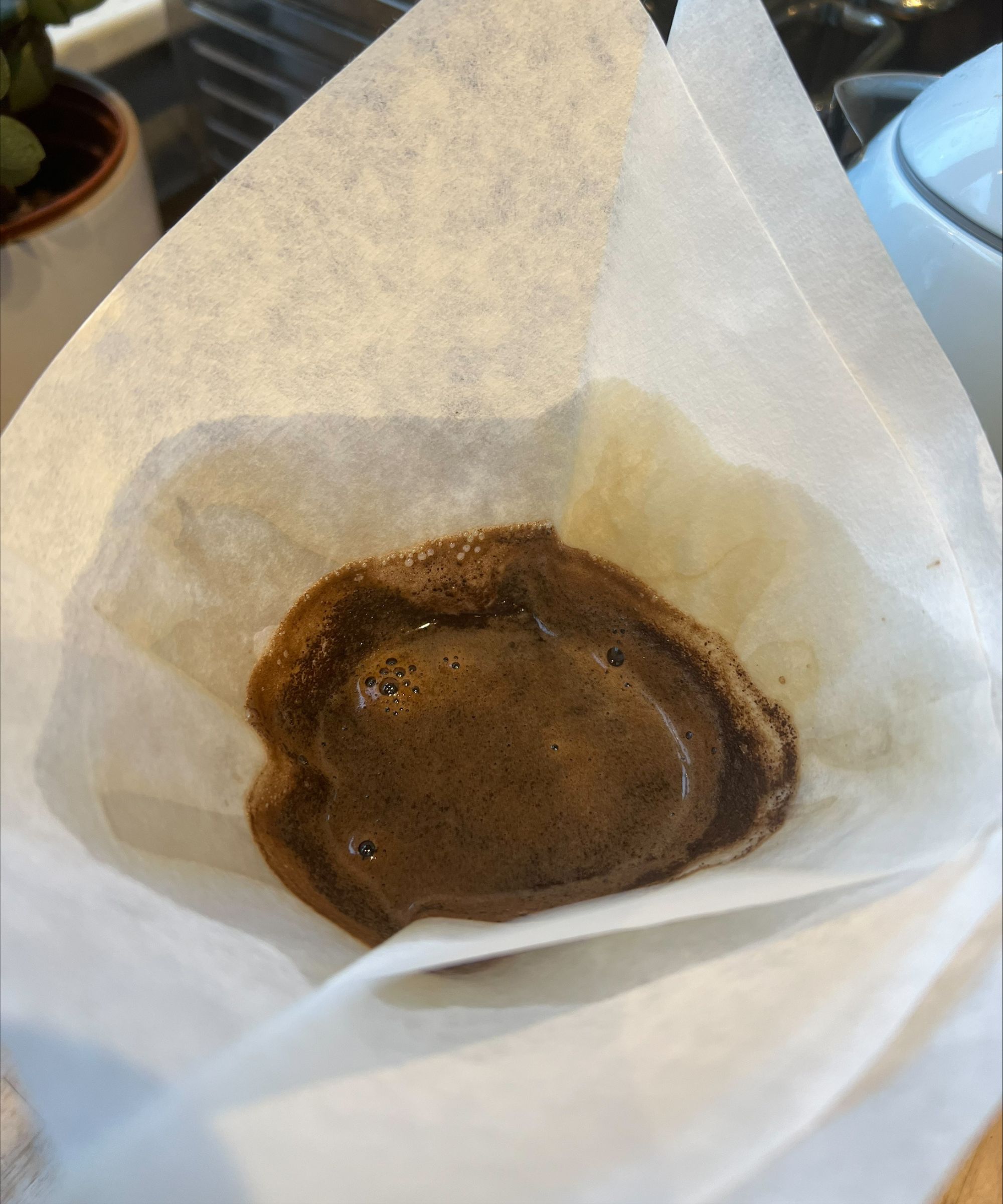 Brewing coffee in the CHEMEX coffee maker using a white filter. To the left, the corner of a white pot with house plant and silver toaster is seen, and to the right is the corner of a baby blue kettle
