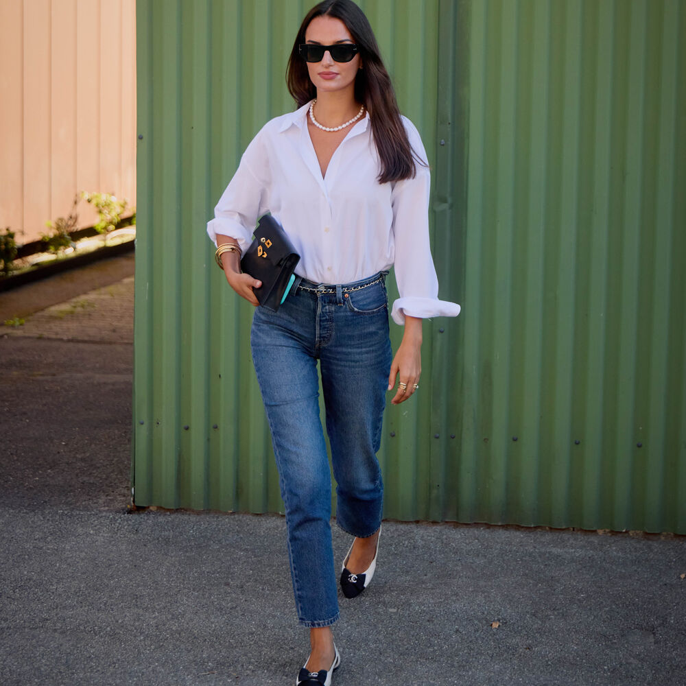 Woman wears button down shirt, jeans, and two toned heels while carrying a black clutch. 