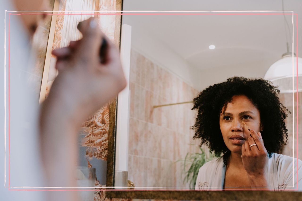 A woman looking in a mirror and applying concealer under her eyes