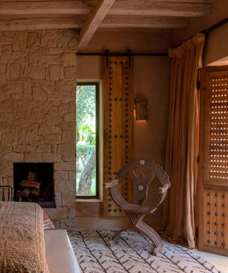 A bedroom with a stone fireplace, a patterned rug, wooden armchair and raftered ceilings