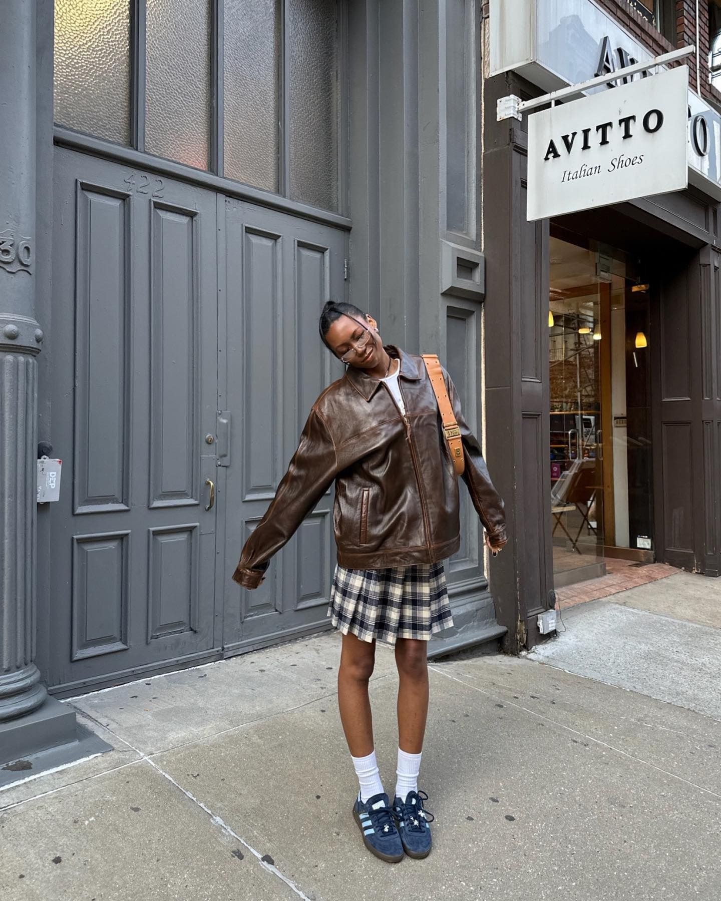 Influencer @chanelmckinsie stands outside a shopfront wearing a brown leather jacket, plaid skirt and navy trainers.