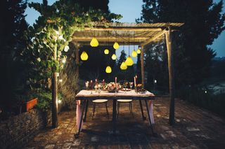 an outdoor pergola in the evening lit by hanging lights, with a dining set and small white stools, with the table full of candles and flowers