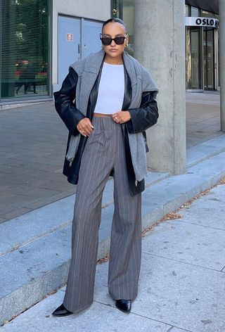 A woman wearing a black leather blazer styled with a white t-shirt, plaid trousers, black pumps, and gray cardigan.