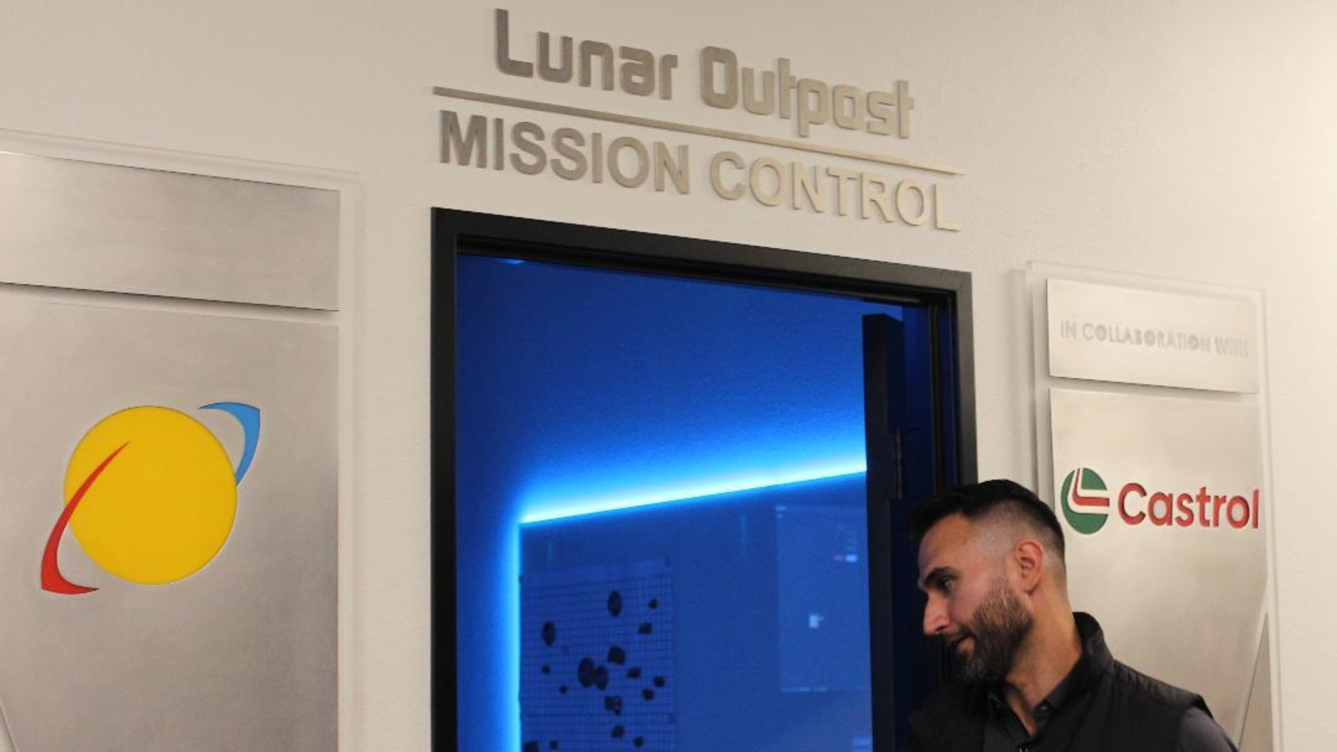 A man wearing a dark polo button up shirt heads into a blue-lit door with the words &quot;Lunar Outpost: Mission Control&quot; above it.