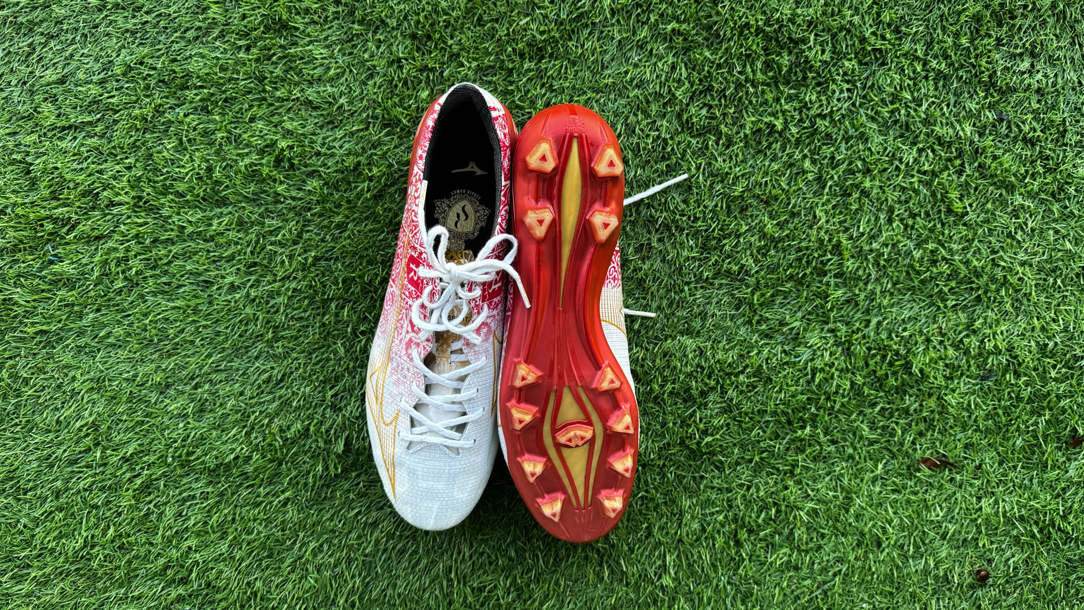 Red and white football boots side by side with one upside down on green artificial grass.