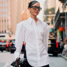 Woman is wearing a white button down top and black skirt while holding a handbag with lots of accessories. She is also wearing sunglasses. 