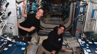 two women astronauts floating on the international space station. one points to an american flag