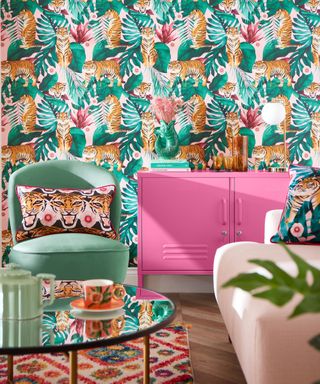 A living area with green and orange leaf and tiger wallpaper, a teal blue chair with a tiger pillow, a pink console table, a light pink couch, and a round glass table