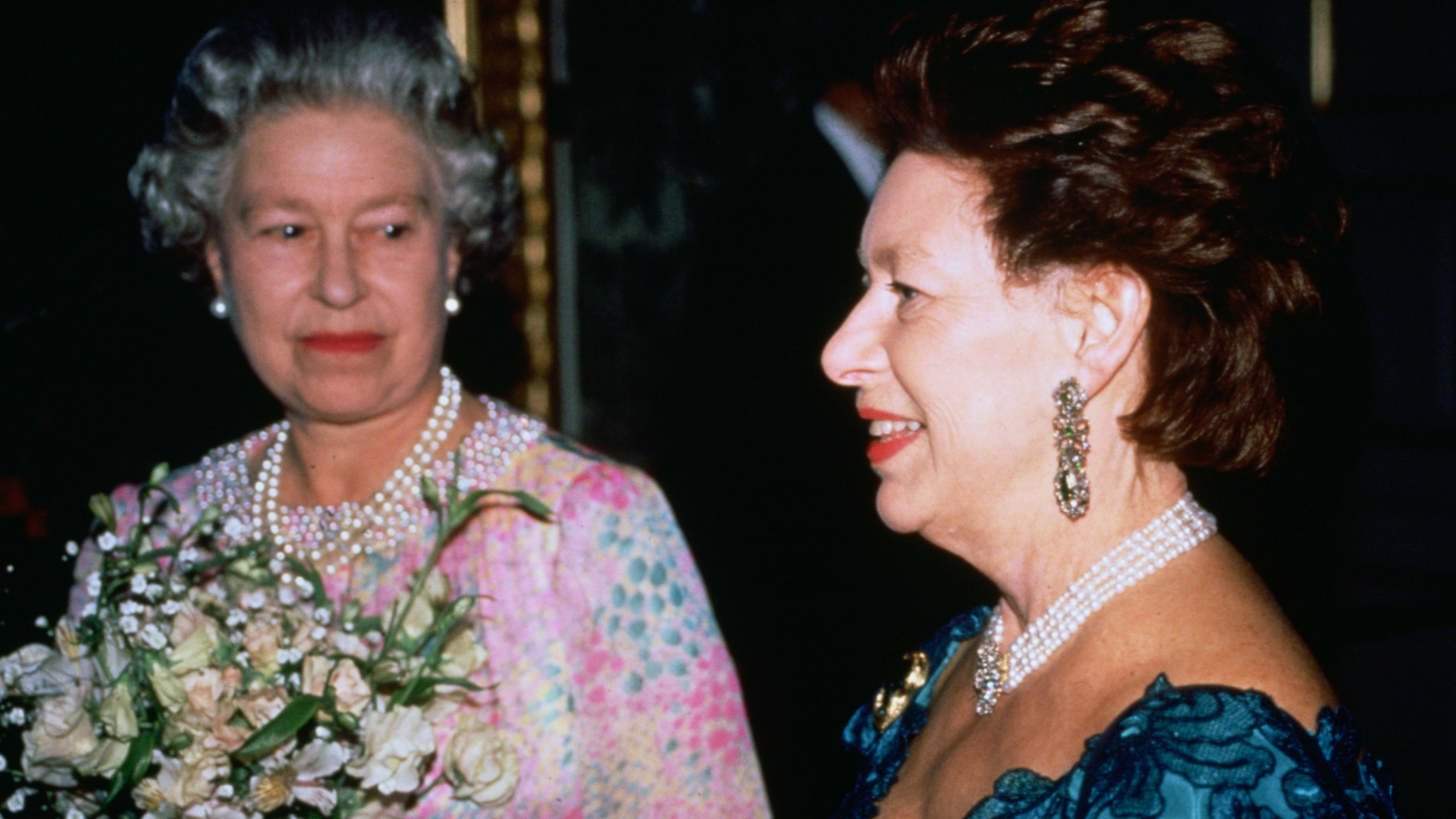 Queen Elizabeth wearing a patterned dress and holding flowers smiling at Princess Margaret