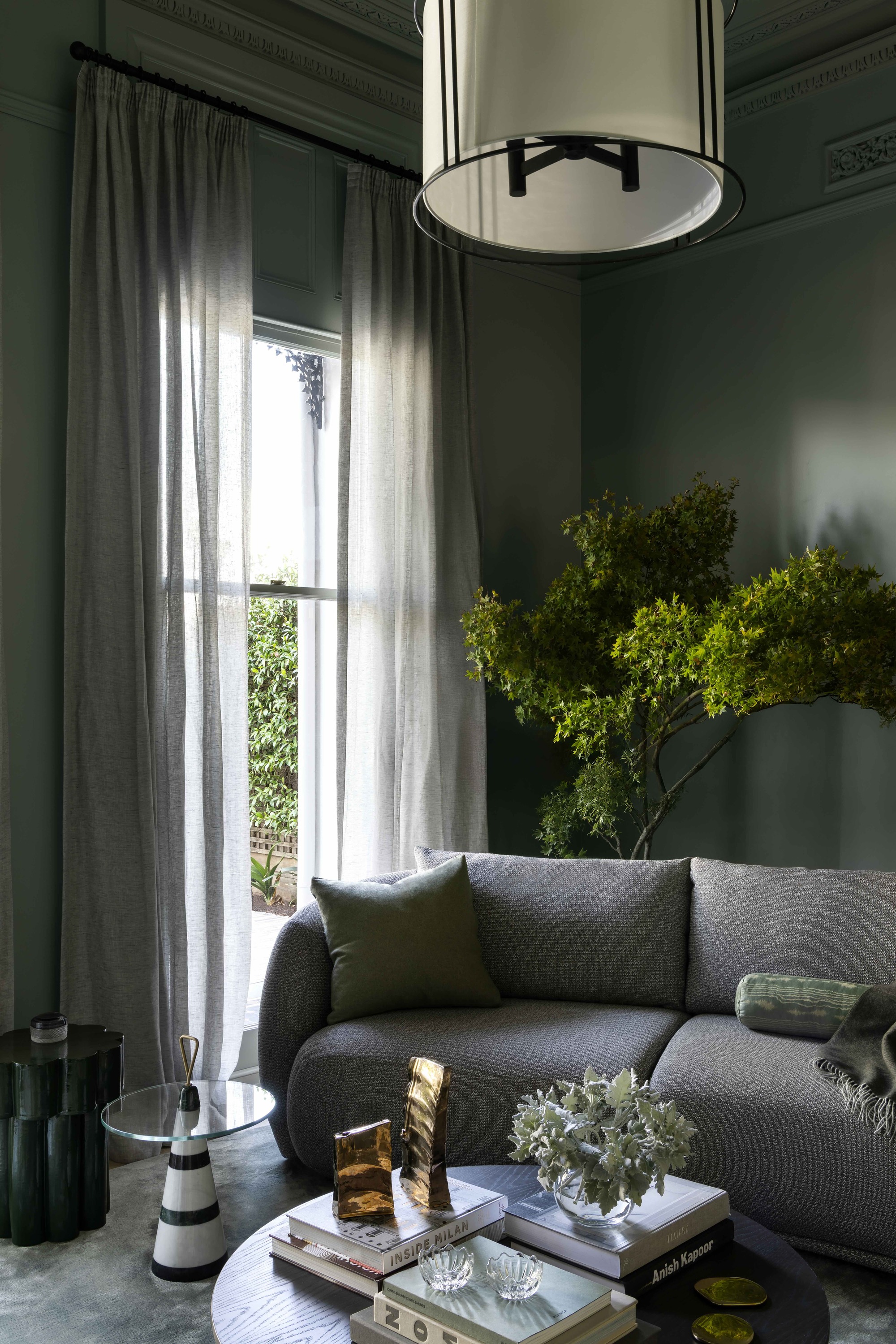 dark gray living room with sheer curtains, gray sofa, round coffee table styled with books and vases, a glass side table, a large plant in the corner of the room behind the sofa