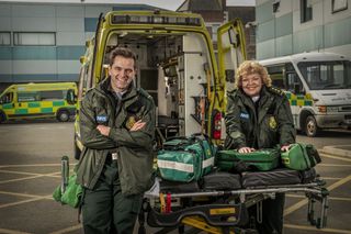 A posed shot of Michael Stevenson and Di Botcher, leaning on the back of an open ambulance in paramedic gear as their characters Iain Dean and Jan Jenning in Casualty.