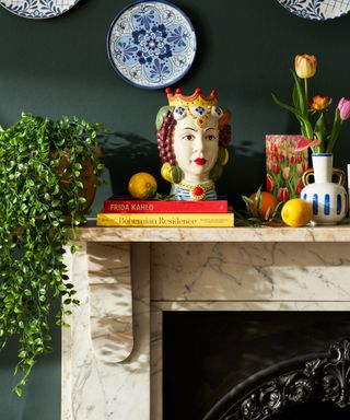 A marble living room fireplace with a mantel with a trailing plant, red and yellow books with a head vase on it and a vase of yellow tulips next to it, against a dark green wall with white and blue plates on it