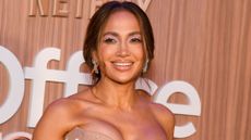 jennifer lopez pictured smiling with a beige dress on a nude-toned backdrop with sparkling earrings, her hair swept back and glamorous makeup