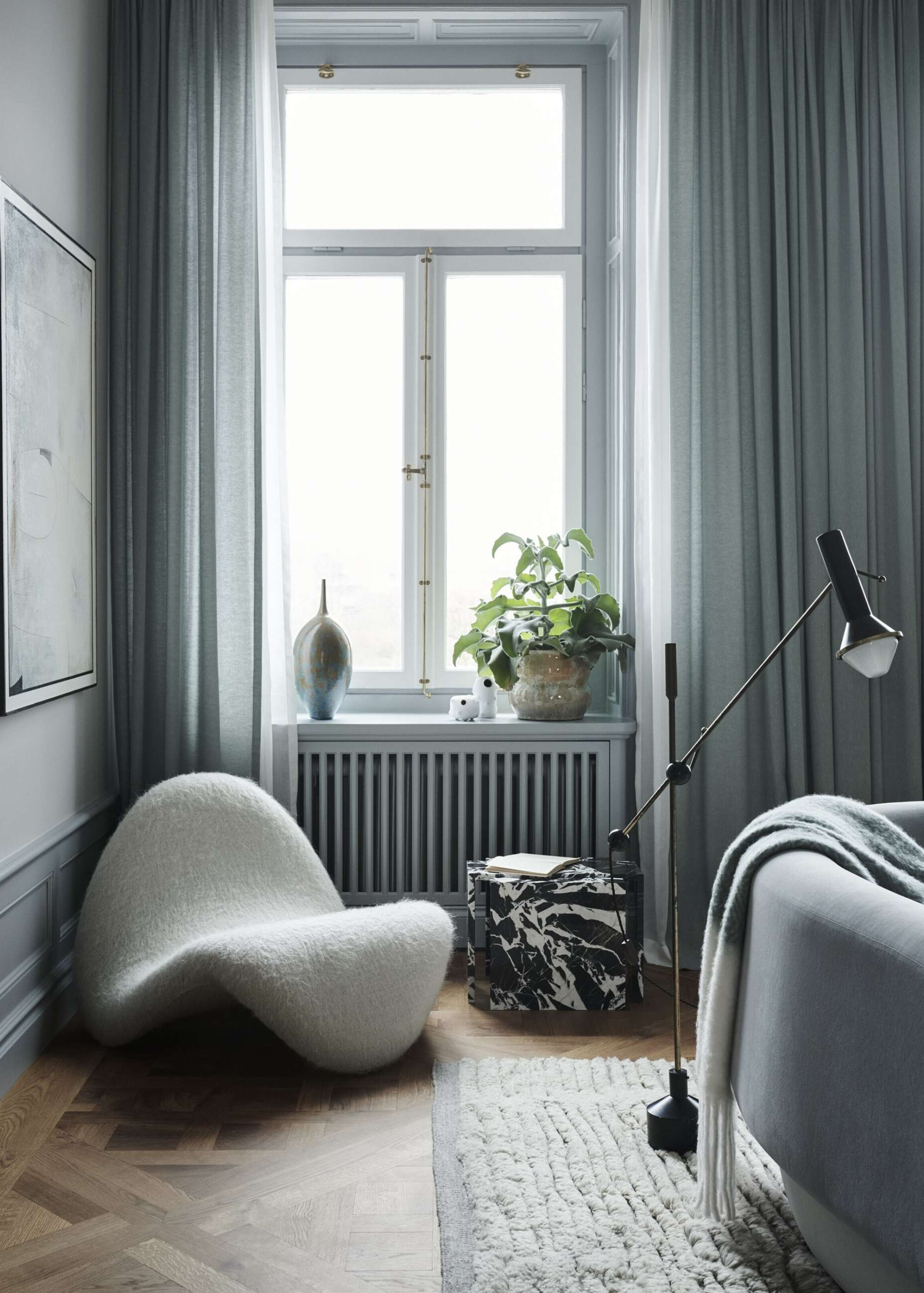 armchair and marble side table styled in a living room next to a window with curtains, a radiator, with a pot plant and ceramic vase on the windowsill. in the foreground, a floor lamp leans over a curved sofa