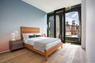 A bedroom with a double bed with a wooden base, a wooden side table with lamps on, a wooden floor and an open sliding door with the view of the city.