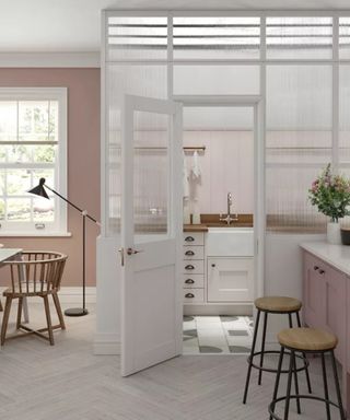 a pink and white utility room idea that is separated from the main kitchen, with a kitchen island and stools to the right, and a dining table to the left