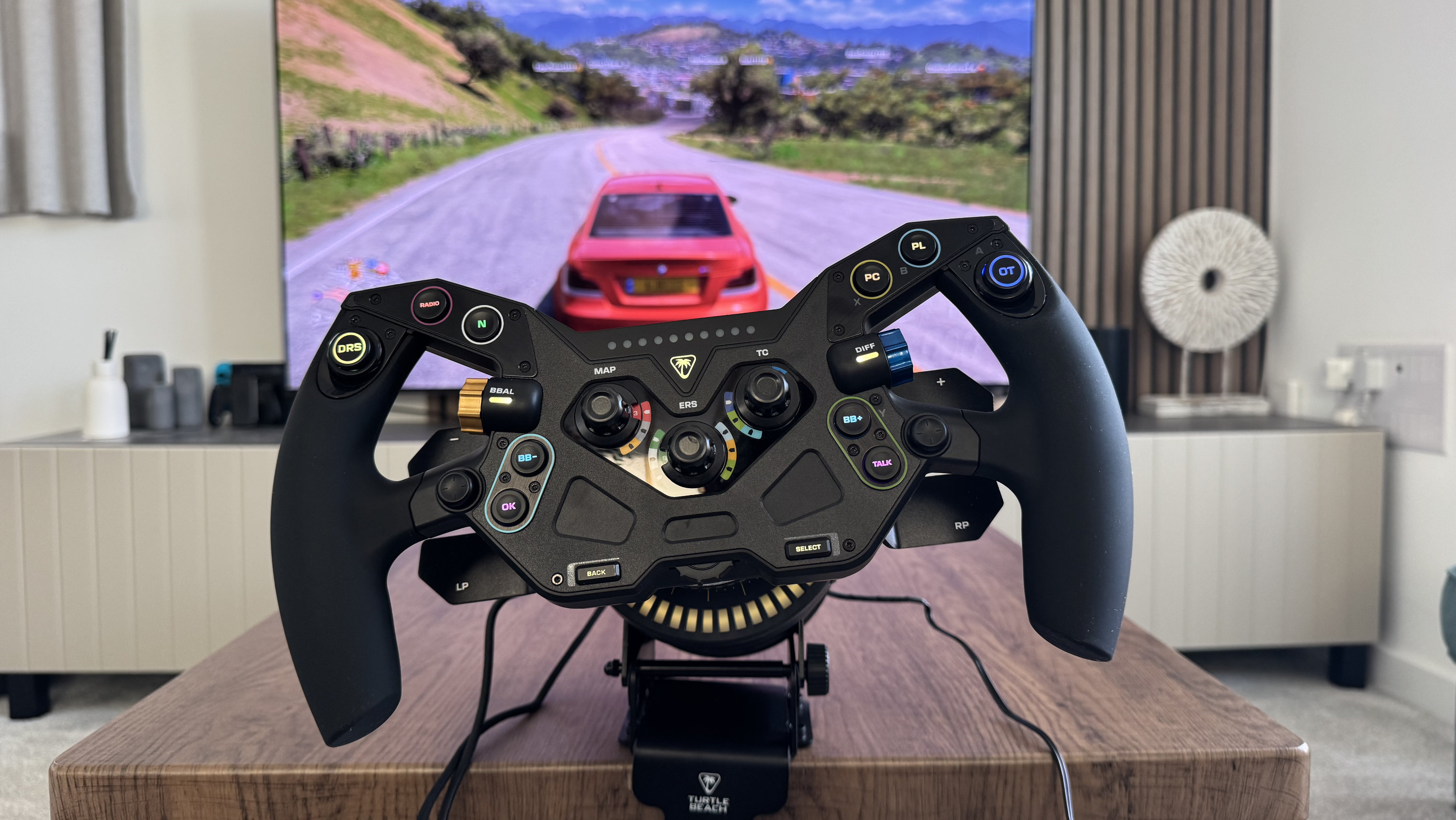The Turtle Beach VelocityOne Race KD3 track wheel mounted to a coffee table, in the background, a racing game on a TV