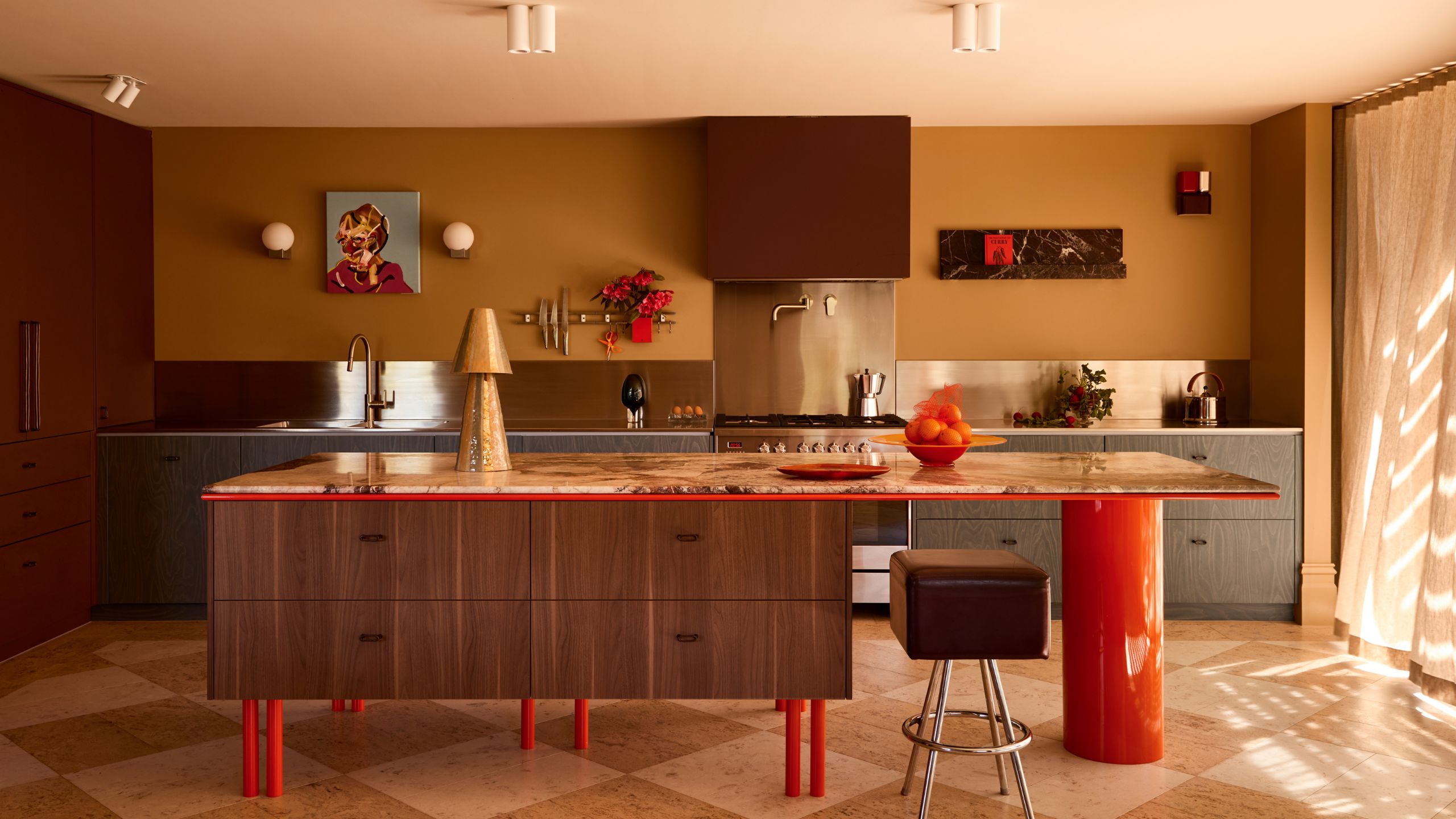 A modern kitchen with a kitchen island with vibrant lacquer red legs and a large circular leg on the seating side, with wood cabinetry and oxblood and blue cabinets against a mustard wall