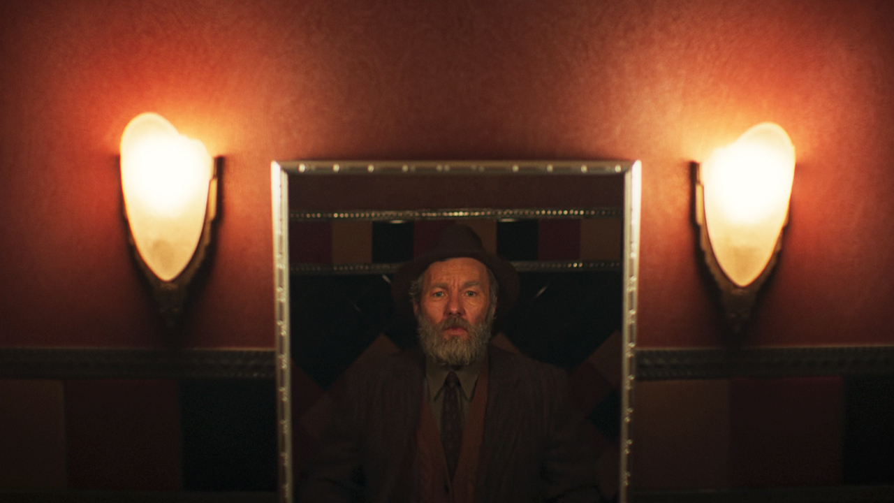 Joel Edgerton looking into a mirror with electric lights on either side in Train Dreams