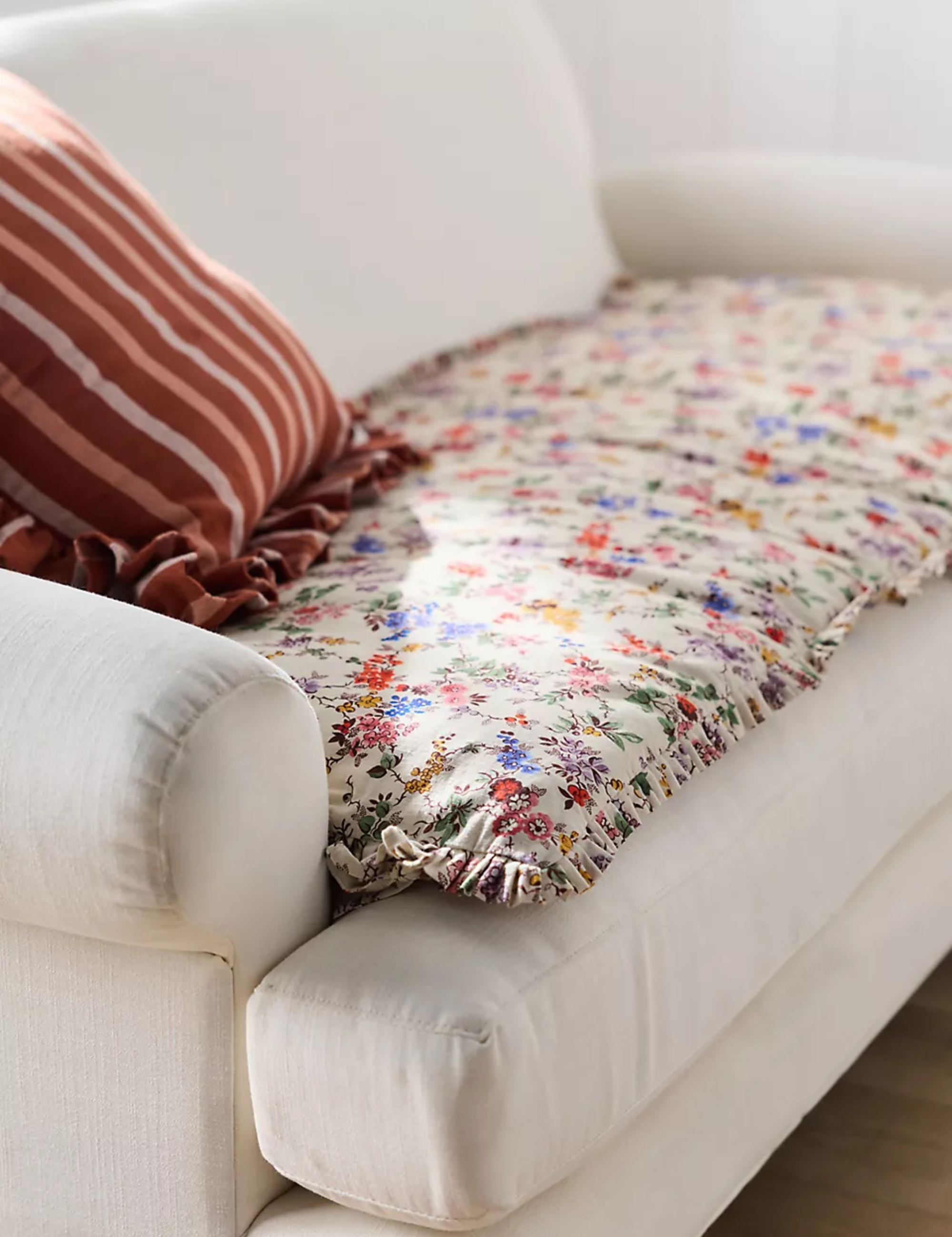 Floral ruffle-bordered Anthropologie sofa topper styled atop of a cream sofa alongside a coordinating striped throw pillow